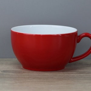 Könnte beinhalten: Eine rote Keramiktasse mit weißem Innenraum. Die Tasse hat eine abgerundete Form und einen Henkel.