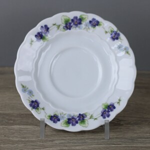 May include: White porcelain saucer with a scalloped edge and a floral design featuring purple and blue violets.