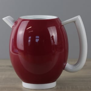 Może przedstawiać: Czerwony ceramiczny czajnik z białym uchwytem i dzióbkiem. Czajnik ma okrągły korpus i białą podstawę.