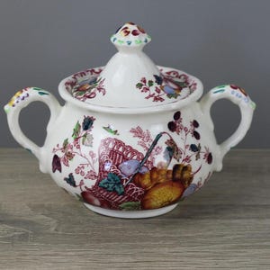 Könnte beinhalten: Eine weiße Zuckerdose aus Keramik mit Deckel, verziert mit einem farbenfrohen Blumen- und Fruchtmuster. Die Dose hat zwei Griffe und einen dekorativen Knopf auf dem Deckel. Das Design umfasst rote, grüne und blaue Akzente.