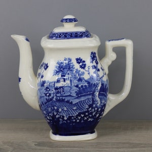 Könnte beinhalten: Eine blaue und weiße Porzellan-Teekanne mit Deckel und Henkel. Die Teekanne zeigt ein blaues und weißes Landschaftsdesign mit einem Haus und Bäumen.