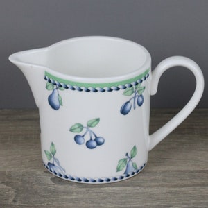 May include: White ceramic creamer with a handle and spout. The creamer is decorated with blue and green fruit illustrations, including pears and cherries. A green and blue border accents the top edge.