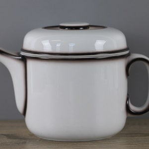 May include: A white ceramic teapot with a rounded body, a curved handle, and a spout. The teapot has a dark brown trim around the lid, spout, and handle. The lid has a small, round knob. The teapot is on a wooden surface.