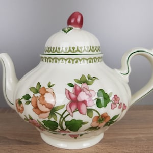 Może przedstawiać: Biały czajnik z kwiatowym wzorem. Czajnik ma zielono-białą obwódkę i różowo-pomarańczowy wzór kwiatowy. Czajnik ma pokrywkę z małym czerwonym uchwytem.