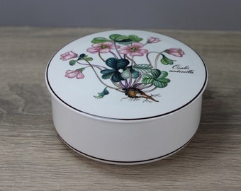 May include: A white ceramic round trinket box with a lid featuring a botanical illustration of pink flowers and green leaves. The box has a black trim and the text "Oxalis acetosella".