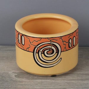 May include: A yellow ceramic planter with a geometric design in black and orange. The planter has a wide, round opening and a smooth, rounded bottom.