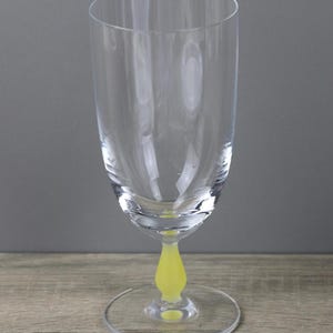 Könnte beinhalten: Ein klares Sektglas mit gelbem Stiel und Fuß. Das Glas hat eine hohe, schlanke Kuppa und einen abgerundeten Fuß. Der Stiel ist leuchtend gelb und der Fuß hat eine klare, kreisförmige Form. Der Hintergrund ist neutral.