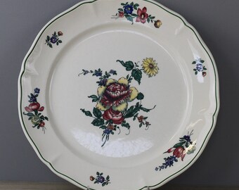 May include: A white ceramic plate with a scalloped edge and a green trim. The plate features a central floral design with yellow, red, and pink flowers, and smaller floral clusters around the rim. The plate is displayed on a clear stand.