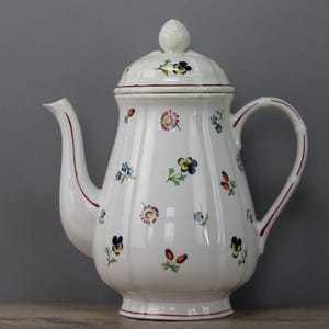 Könnte beinhalten: Weiße Keramik-Teekanne mit Deckel, verziert mit bunten Blumenmustern. Die Teekanne hat einen gebogenen Ausguss, einen abgerundeten Griff und einen dekorativen Knauf auf dem Deckel. Rote Verzierungen akzentuieren Ausguss und Griff.