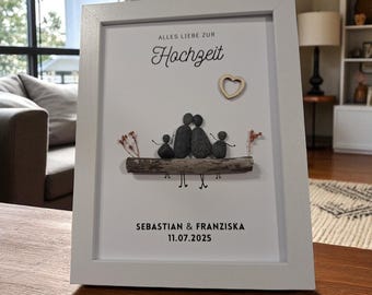 May include: A framed art piece with a white border. The artwork features a pebble art family on a wooden base, with the text "ALLES LIEBE ZUR Hochzeit" and names "SEBASTIAN & FRANZISKA" with the date "11.07.2025". A wooden heart is also included.