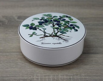 May include: A round, off-white ceramic container with a lid featuring a botanical illustration of a blueberry plant. The illustration includes green leaves, dark blue berries, and the text "Vaccinium myrtillus". The container has a black trim.