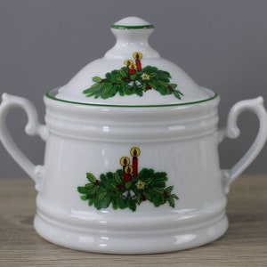 May include: White ceramic sugar bowl with a lid, decorated with a Christmas wreath design including candles, bells, and stars. The bowl has two handles and a green trim. A festive piece for serving sugar or other condiments.