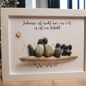 Könnte beinhalten: Ein gerahmtes Kunstwerk mit einer Familie aus fünf Kieselsteinen, die sich an einem Stück Treibholz die Hände halten. Das Kunstwerk hat einen weißen Hintergrund und einen weißen Rahmen. Der Text "Zuhause ist nicht nur ein Ort. es ist ein Gefühl." ist in schwarzer Kursivschrift geschrieben.
