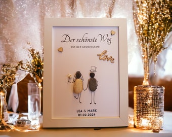 May include: A white framed sign with the text "Der schönste Weg ist der gemeinsame! Love" and a drawing of a couple made of stones. The sign also includes the names "Lea & Mark" and the date "01.02.2024".