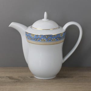 Può includere: Teiera in ceramica bianca con coperchio e manico. La teiera presenta una fascia floreale blu e oro al centro e finiture dorate. Il beccuccio è sul lato sinistro e il manico sul lato destro.