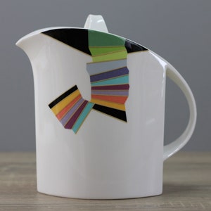 Peut inclure: Pichet en céramique blanche avec un motif géométrique en noir, vert, jaune, orange, violet et bleu. Le motif est une série de rayures qui forment une forme incurvée.
