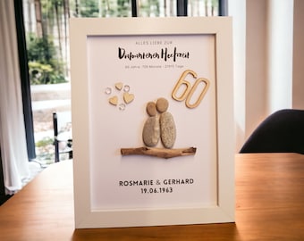 May include: A framed artwork celebrating a 60th wedding anniversary. The white framed art features a pebble art couple, wooden hearts, and the number 60. Text includes the couple's names and anniversary date. The artwork is displayed on a wooden table.