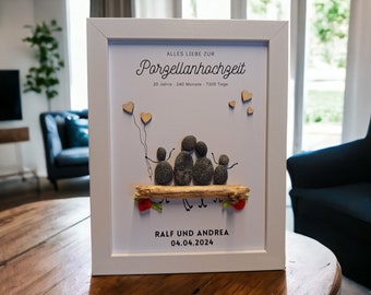 May include: A white framed picture with a white background. The picture features a family of five made of gray stones sitting on a piece of driftwood. The stones are holding hands and have small stick arms and legs. There are small wooden hearts above the family. The text "ALLES LIEBE ZUR Porzellanhochzeit 20 Jahre 240 Monate 7300 Tage" is at the top of the picture. The text "RALF UND ANDREA 04.04.2024" is at the bottom of the picture.