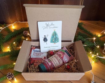 May include: A Christmas gift box with a greeting card, a tube of cream, and a package of tea. The card has a drawing of a Christmas tree and the words "Wishing you a wonderful Christmas!" The box is decorated with pine branches and lights.