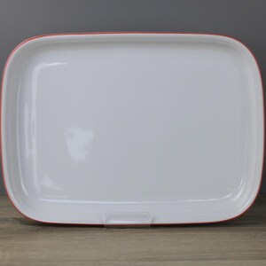 Peut inclure: Plateau de service rectangulaire blanc avec un bord rouge. Le plateau a des coins arrondis et une petite poignée sur un côté. Le plateau est vide et repose sur une surface en bois.