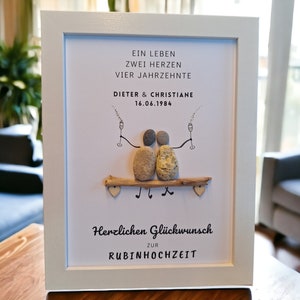 Könnte beinhalten: Ein weiß gerahmtes Bild mit weißem Hintergrund. Das Bild zeigt ein Paar, das mit Kieselsteinen dargestellt ist, auf einem Ast sitzt und Champagnergläser hält. Der Text lautet "Ein Leben Zwei Herzen Vier Jahrzehnte Dieter & Christiane 16.06.1984 Herzlichen Glückwunsch Zur Rubinhochzeit".