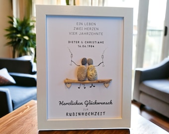 May include: A white framed picture with a white background. The picture features a couple depicted with pebble stones, sitting on a branch, holding glasses of champagne. The text reads "Ein Leben Zwei Herzen Vier Jahrzehnte Dieter & Christiane 16.06.1984 Herzlichen Glückwunsch Zur Rubinhochzeit".
