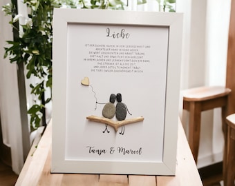 May include: A white framed print with a gray background and a quote about love. The quote reads, "Liebe ist der sichere Hafen, in dem Geborgenheut und Abenteuer Hand in Hand gehen..." The print also features a small, brown heart with a string attached to it, and two small, gray figures holding hands. The figures are sitting on a small, brown branch. The print is signed "Tanja & Marcel".