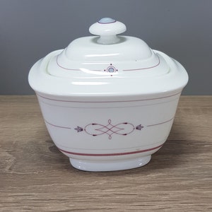 May include: White ceramic sugar bowl with a lid and a maroon design. The bowl has a rounded shape with a small base and a matching lid with a small knob. The maroon design includes lines and a central flourish.