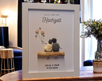 May include: A white framed picture with a white background. The picture features a couple made of stones sitting on a piece of driftwood. The couple is holding balloons in the shape of hearts. The text "Alles Liebe zur Hochzeit" is at the top of the picture. The text "Julia & Max 17.05.2024" is at the bottom of the picture.