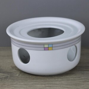 Könnte beinhalten: Weißer Keramik-Teewärmer mit einer kreisförmigen Öffnung und zwei seitlichen Öffnungen. Der Wärmer hat ein graues Band mit einem farbenfrohen quadratischen Muster. Das Design ist schlicht und elegant, für den Hausgebrauch geeignet.