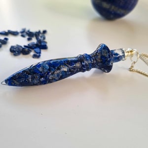 May include: A blue lapis lazuli pendulum with a silver chain. The pendulum is made of many small pieces of lapis lazuli that have been glued together.