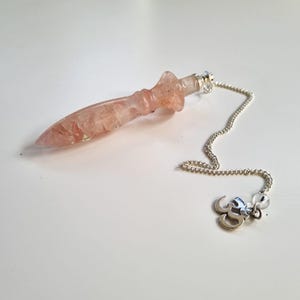 Può includere: Un pendolo di cristallo rosa con una catena d'argento e un ciondolo decorativo. Il cristallo ha un'estremità appuntita e un manico arrotondato. La catena è attaccata al cristallo con un piccolo cappuccio d'argento. Il ciondolo è un simbolo d'argento.