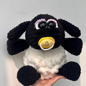 Puede incluir: Un juguete de oveja de ganchillo hecho a mano. Tiene cabeza, orejas y extremidades negras, y un cuerpo blanco. La oveja tiene ojos negros grandes, un chupete amarillo y una ceja rosa. El juguete es suave y afelpado, para niños.