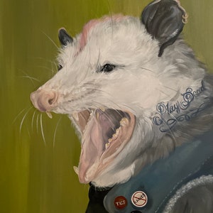 May include: A white opossum with pink ears and a black nose is wearing a blue denim jacket with patches. The opossum has a tattoo that says "Play Dead" on its side. The opossum is yawning with its mouth open, showing its teeth.