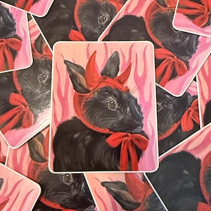 May include: A sticker featuring a black rabbit with red devil horns and a red bow. The rabbit is set against a pink background with a flame-like design. The sticker has a white border.