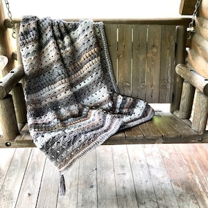 May include: A crocheted throw blanket in shades of brown, beige, and white is draped over a wooden porch swing. The blanket has a fringe detail and a textured pattern.