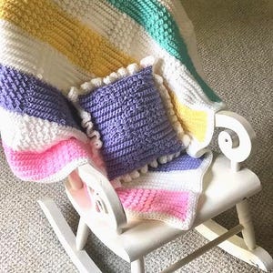May include: A white wooden rocking chair with a crocheted blanket and pillow. The blanket features stripes of yellow, white, mint green, and pink. A small purple pillow with a ruffled edge rests on the chair.