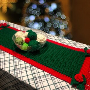 May include: A green crocheted table runner with a red border and red and green crocheted flowers. The runner is on a white and green plaid tablecloth. There are balls of yarn in a glass bowl on the runner.