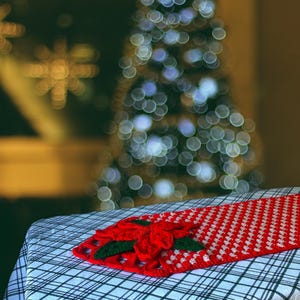 May include: A red and white crocheted table runner with a poinsettia design. The runner is on a black and white checked tablecloth. A blurred Christmas tree with white lights is in the background.