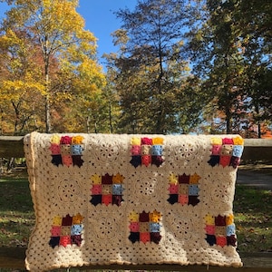 May include: A crocheted blanket with a cream background and colorful squares. The squares are made up of different colored yarn in shades of red, yellow, blue, and pink. The blanket is hanging on a wooden fence with a blurred background of trees.