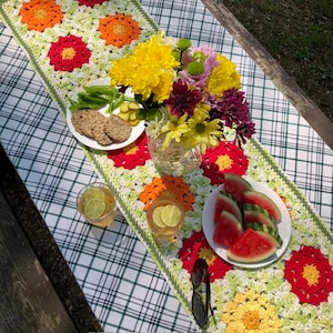 May include: A crocheted table runner with a floral design in shades of red, orange, and yellow. The runner is laid on a checkered tablecloth and features a vase of flowers, watermelon slices, and two glasses of iced tea.