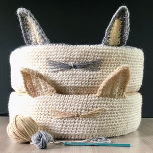 May include: Two crocheted cat-shaped baskets, one stacked on top of the other. The top basket is gray with white accents and the bottom basket is beige with brown accents. Both baskets have crocheted ears and whiskers.