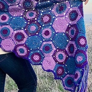 May include: A crocheted shawl with a hexagonal pattern in shades of purple, blue, and white. The shawl has a fringe border and is draped over a person's shoulders.