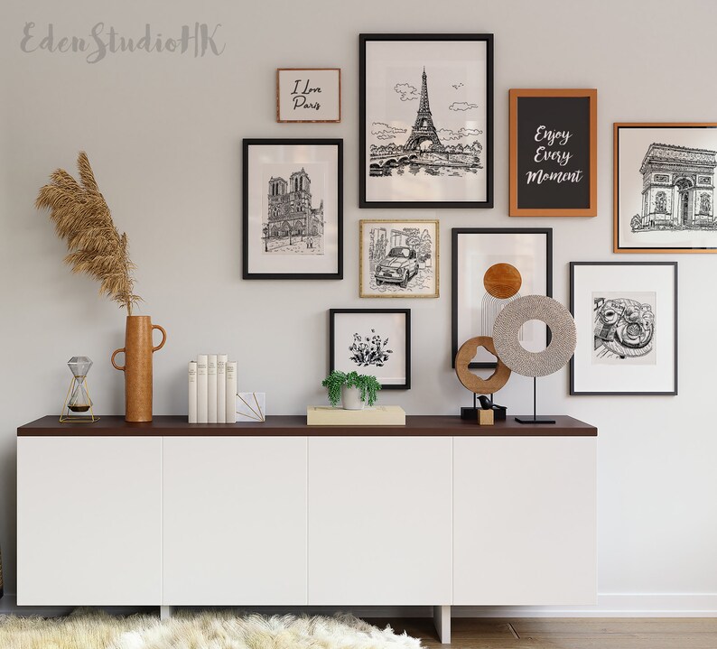 May include: A white cabinet with three doors sits on a dark wood surface. Above the cabinet is a gallery wall with black and white drawings of Paris landmarks, including the Eiffel Tower, Notre Dame Cathedral, and the Arc de Triomphe. The wall also features a wooden sign that says "Enjoy Every Moment".