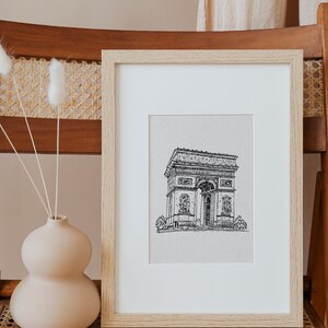 May include: A framed black and white line drawing of the Arc de Triomphe in Paris, France. The drawing is on a white background and is framed in a light wood frame.