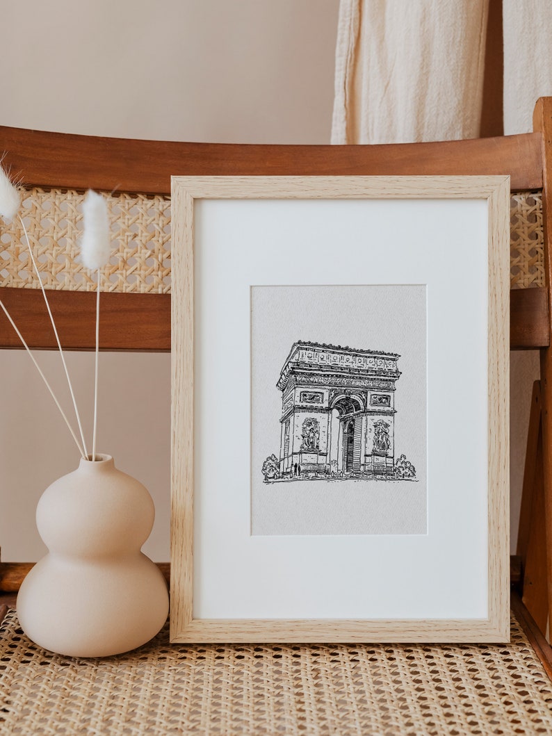 May include: A framed black and white line drawing of the Arc de Triomphe in Paris, France. The drawing is on a white background and is framed in a light wood frame.
