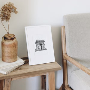 May include: A black and white line drawing of the Arc de Triomphe in Paris, France, framed in a white frame and displayed on a wooden table.