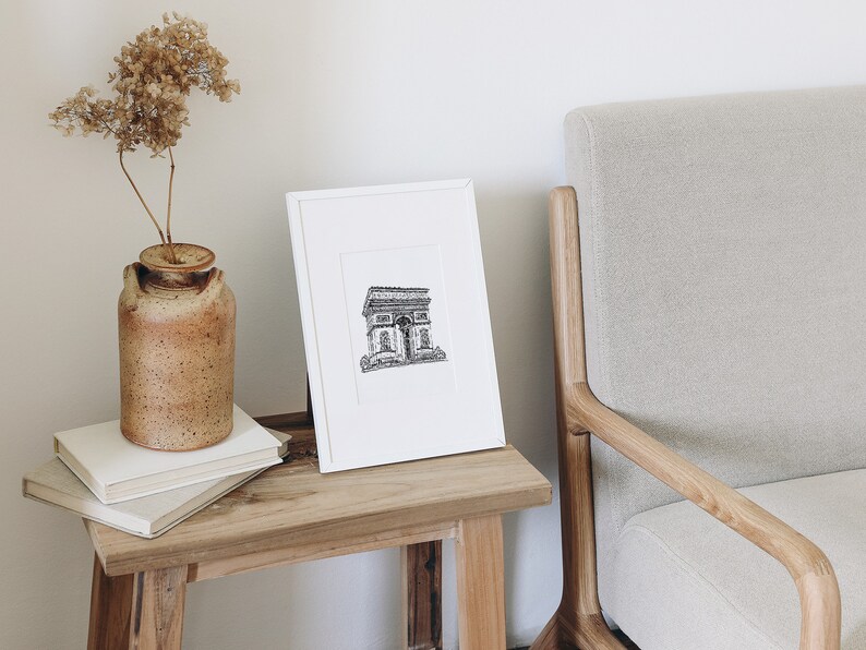 May include: A black and white line drawing of the Arc de Triomphe in Paris, France, framed in a white frame and displayed on a wooden table.