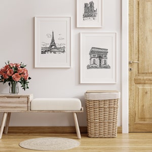 May include: A white wooden bench with a white cushion sits in front of a white wall with three black and white framed prints of Parisian landmarks. A wicker laundry basket sits to the right of the bench. A vase of pink roses sits on a wooden table to the left of the bench.
