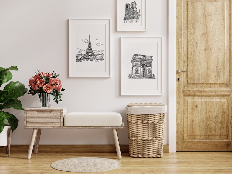 May include: A white wooden bench with a white cushion sits in front of a white wall with three black and white framed prints of Parisian landmarks. A wicker laundry basket sits to the right of the bench. A vase of pink roses sits on a wooden table to the left of the bench.